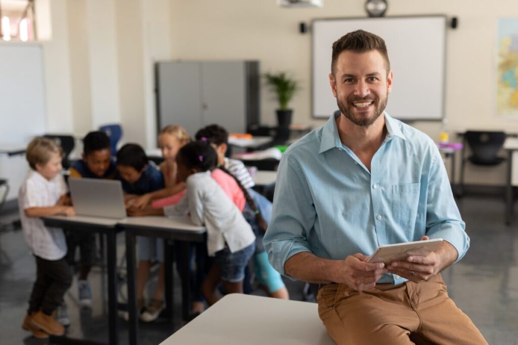 Teacher with digital tablet looking at camera Ein Lehrer sitzt mit seinem Tablett im Klassenraum, hinter ihm arbeiten seine Schüler.