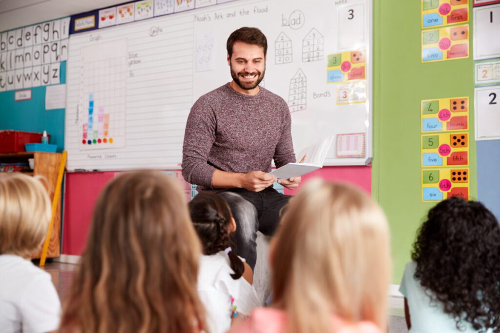 Male Teacher Reading Story To Group Of Elementary Pupils In School Classroom Ein Lehrer sitzt im Klassenraum vor seinen Schülern