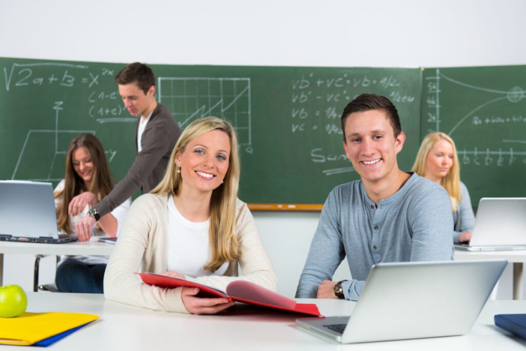 zusammenarbeit im team Eine Lehrerin und ein Schüler sitzen lächelnd im Klassenraum vor einer Tafel.