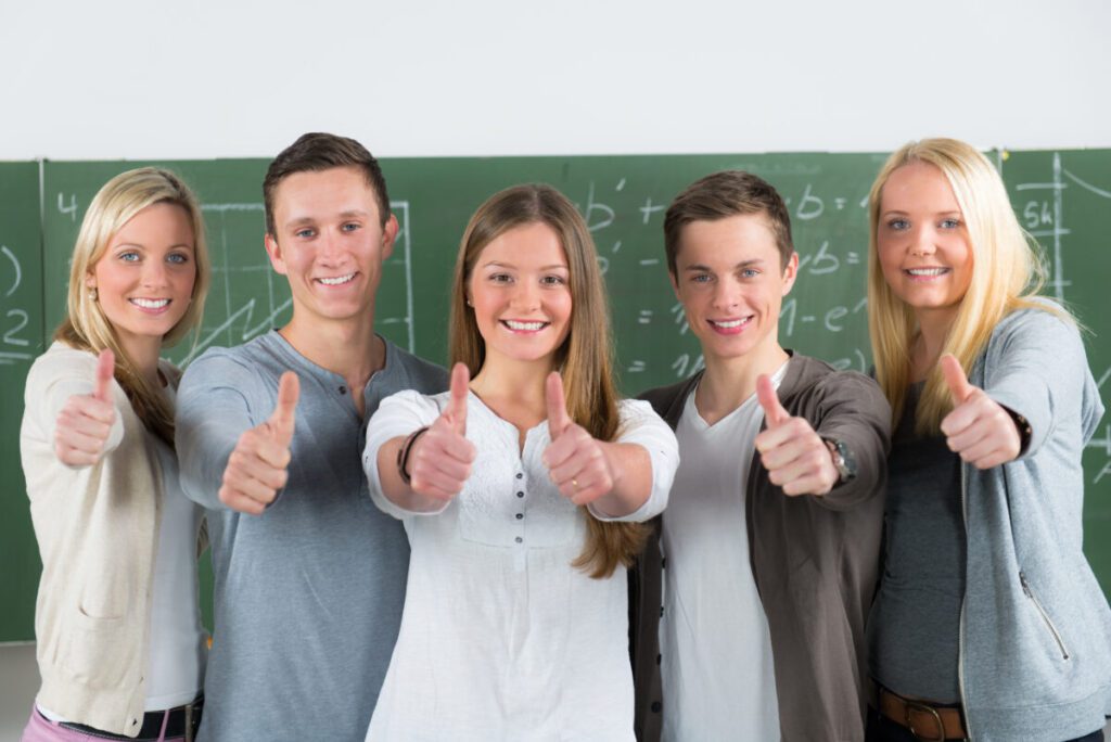 schülerteam zeigt motiviert daumen hoch Schüler stehen in einem Klassenraum vor einer Tafel, lächeln und zeigen den Daumen nach oben