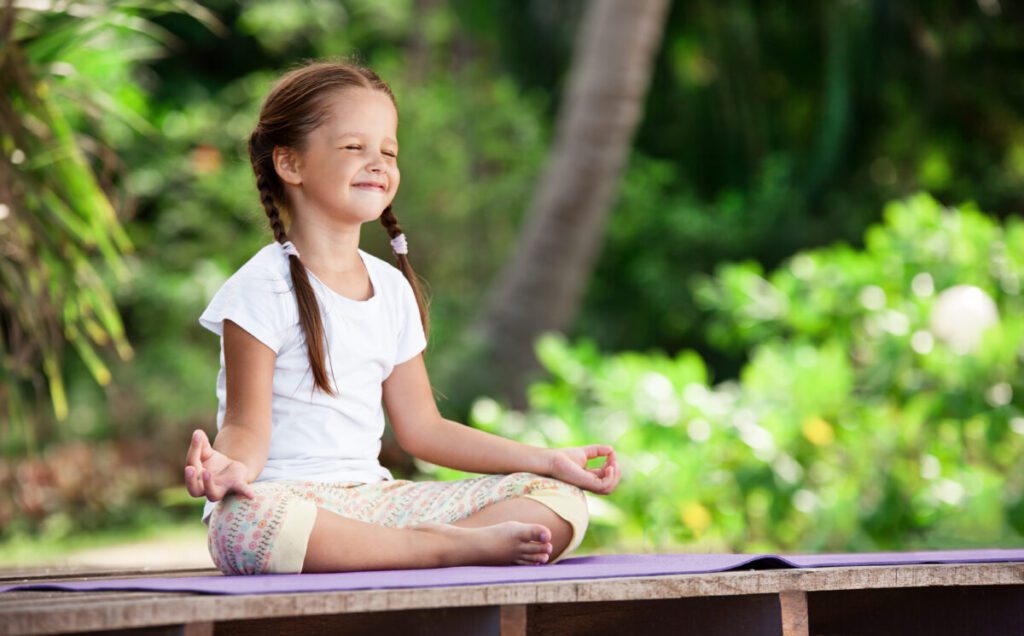 Child doing exercise on platform outdoors. Healthy lifestyle. Yoga girl Ein Mädchen sitzt mit geschlossenen Augen im Grünen auf einer Matte im Schneidersitz und meditiert