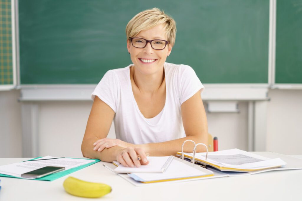 lehrerin bereitet unterricht vor Eine zufriedene, glückliche Lehrerin sitzt am Lehrertisch vor einer Tafel im Klassenraum und lächelt.